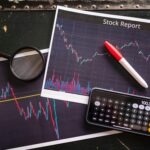 Printed stock charts with candlestick data, a red pen, magnifying glass, calculator and smartphone nearby.