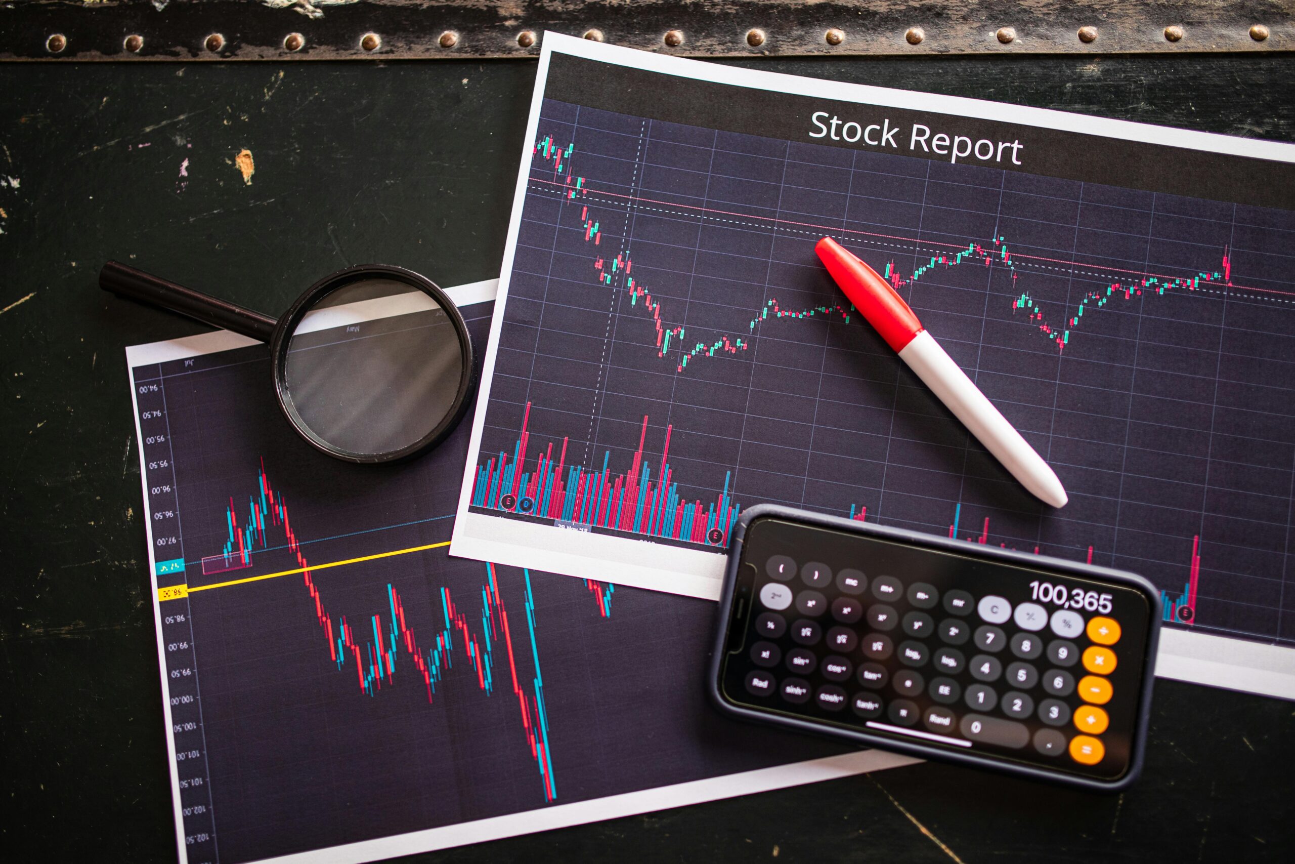 Printed stock charts with candlestick data, a red pen, magnifying glass, calculator and smartphone nearby.