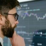 Man with glasses and beard studying a large candlestick stock chart on a monitor.