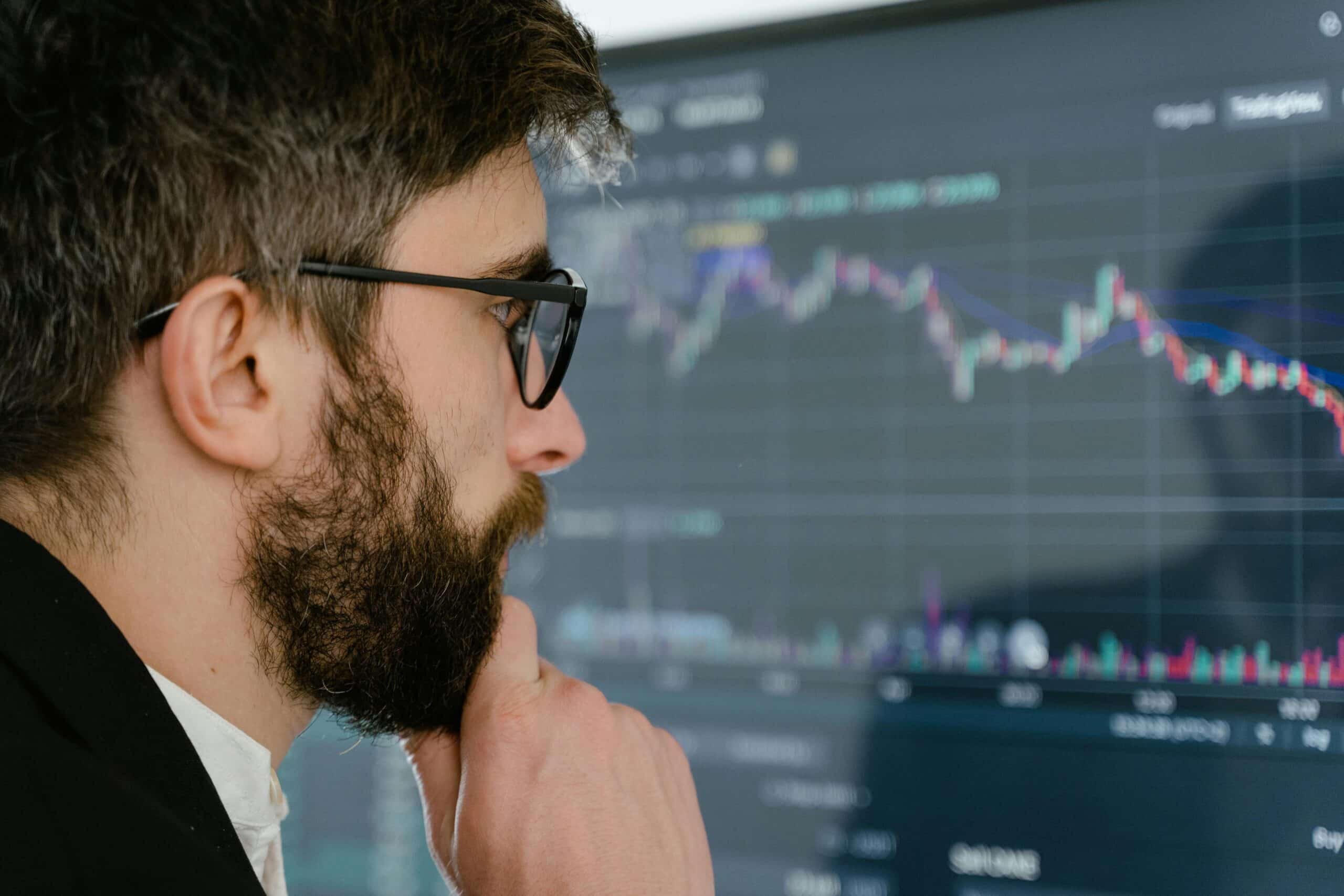 Man with glasses and beard studying a large candlestick stock chart on a monitor.